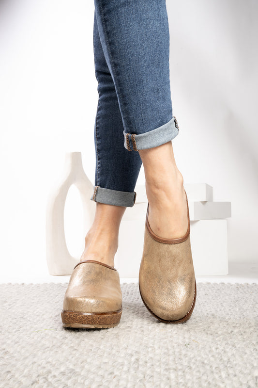 A person wearing blue cuffed jeans and a pair of distressed metallic gold slip-on mules on a light gray textured rug, with a white background and minimalist white decor.