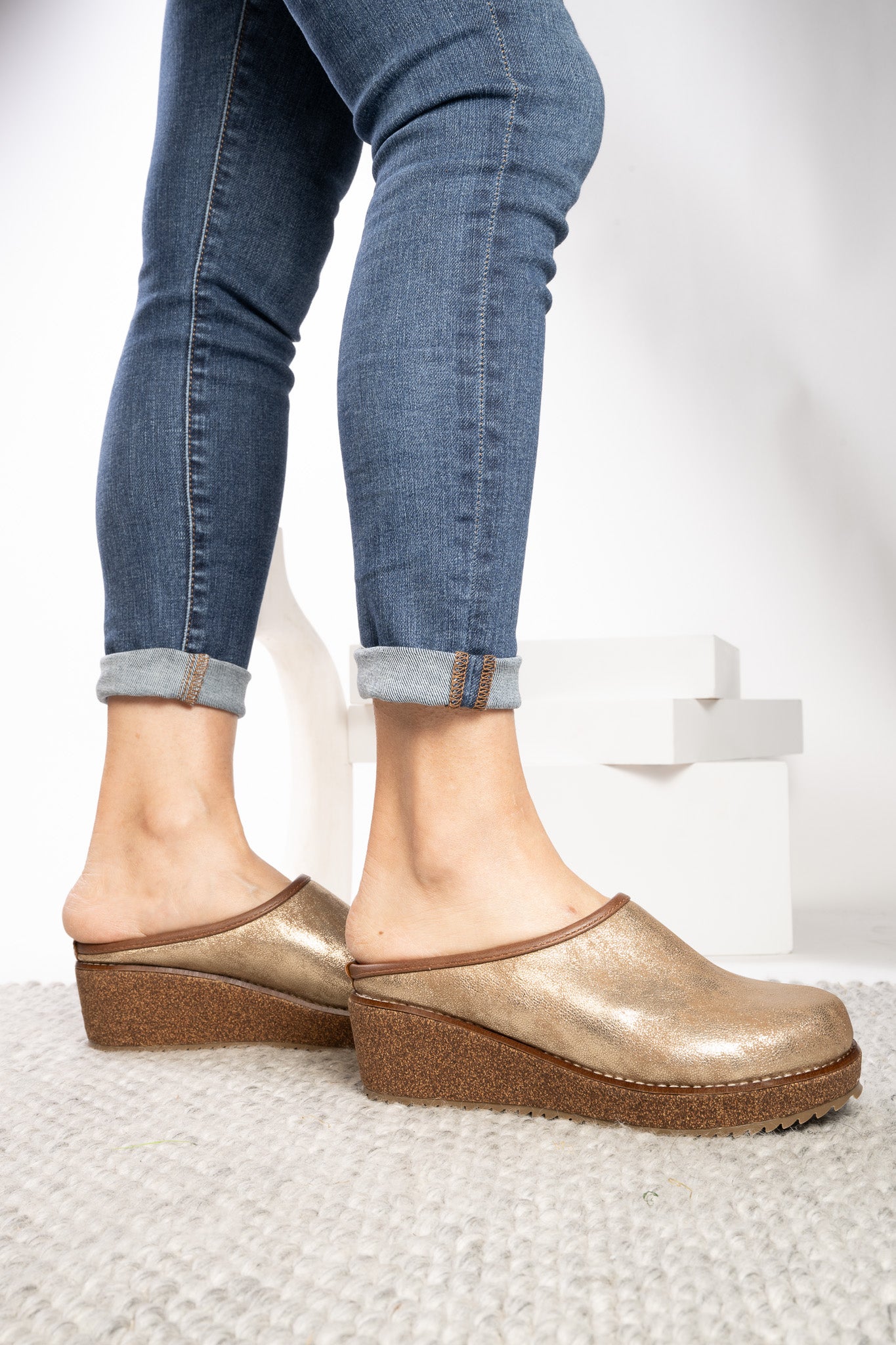 Metallic gold clog-style mule shoes with brown wedge soles, shown on a model wearing blue cuffed jeans against a clean white studio background.