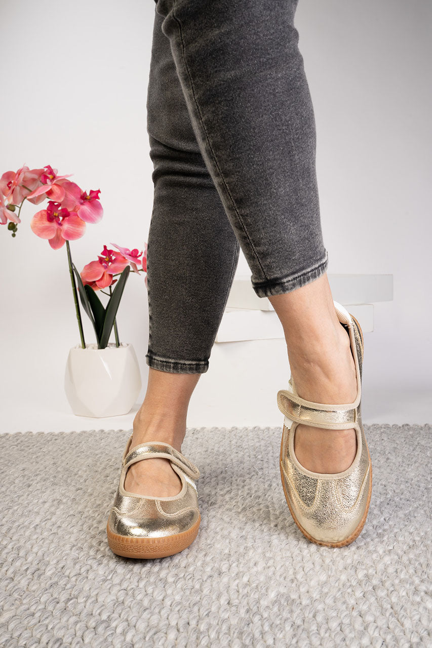 A person wearing dark gray jeans and metallic gold Mary Jane style shoes, standing on a textured light gray rug with a white vase of pink flowers in the background.