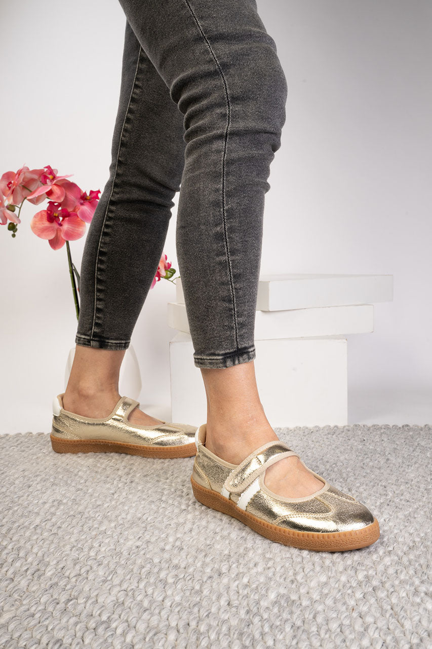 Metallic gold Mary Jane flat shoes worn with dark gray skinny jeans, shown on a model standing on a textured gray rug.