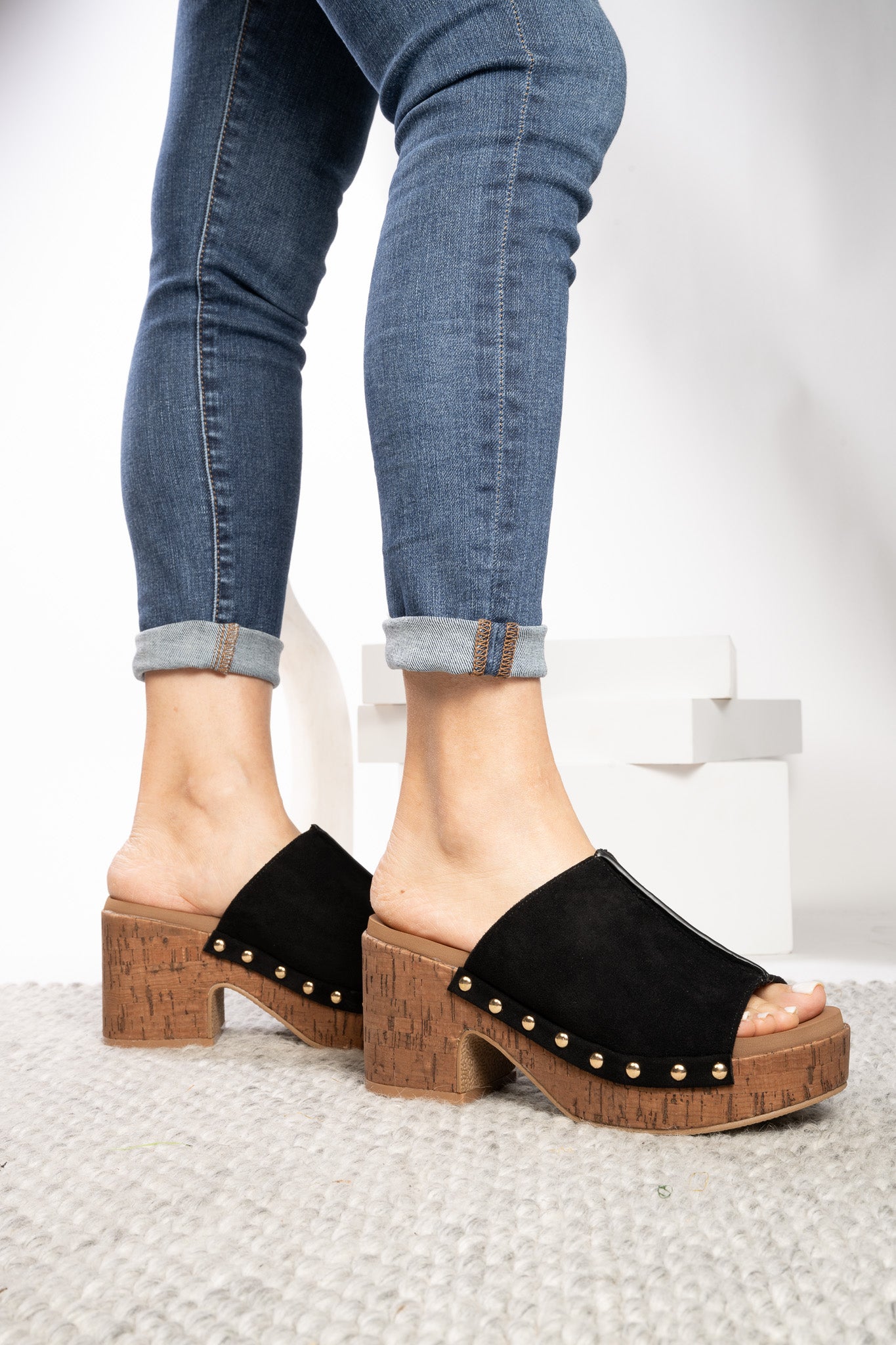 Black suede mule sandals with cork-look block heels and gold-tone studs, shown on a model wearing blue jeans against a white studio background.