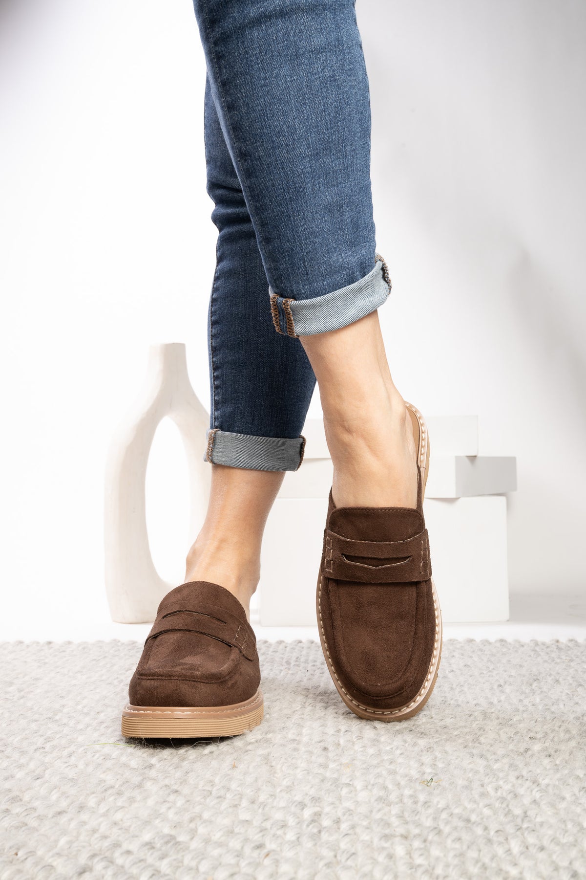 A person wearing brown suede penny loafers with cuffed blue jeans, standing on a light gray textured rug in a bright studio setting.