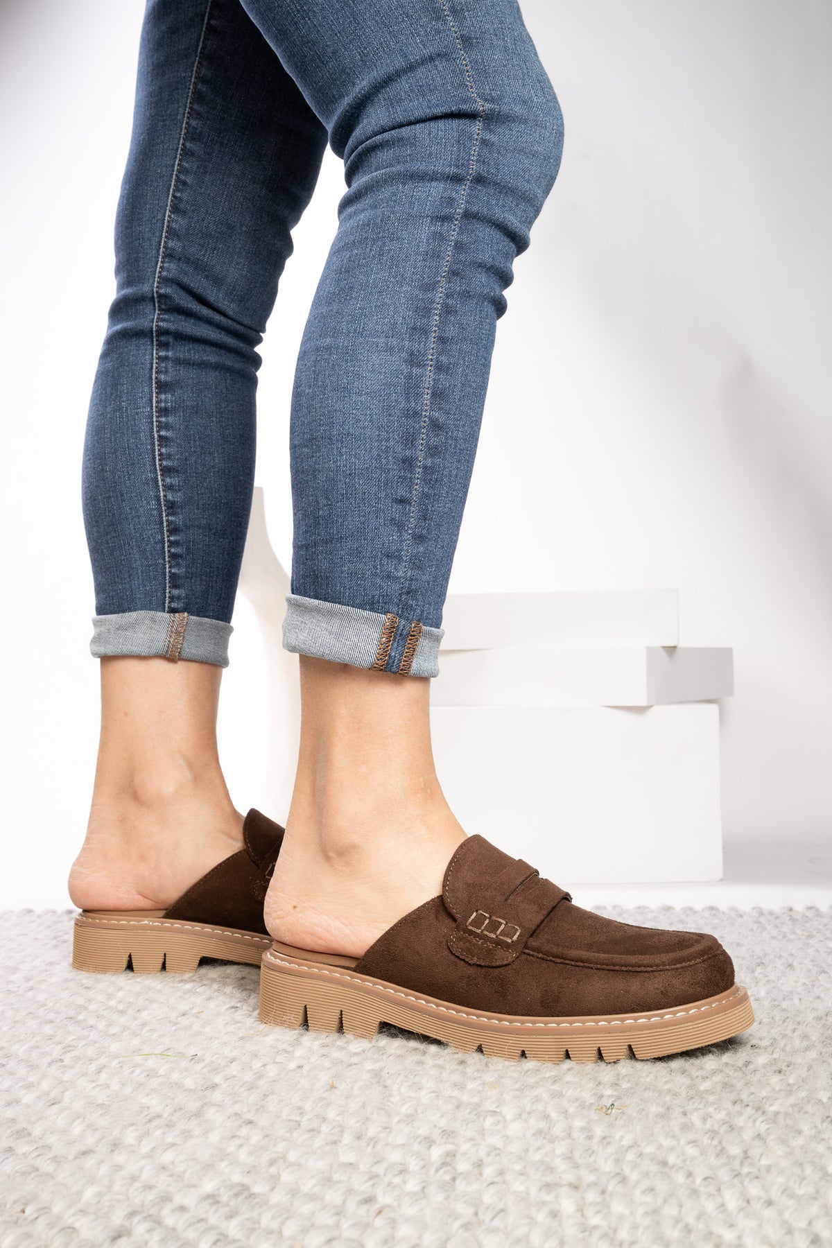 A person wearing brown suede mule loafers with chunky beige soles and rolled-up blue jeans, standing on a light textured rug.