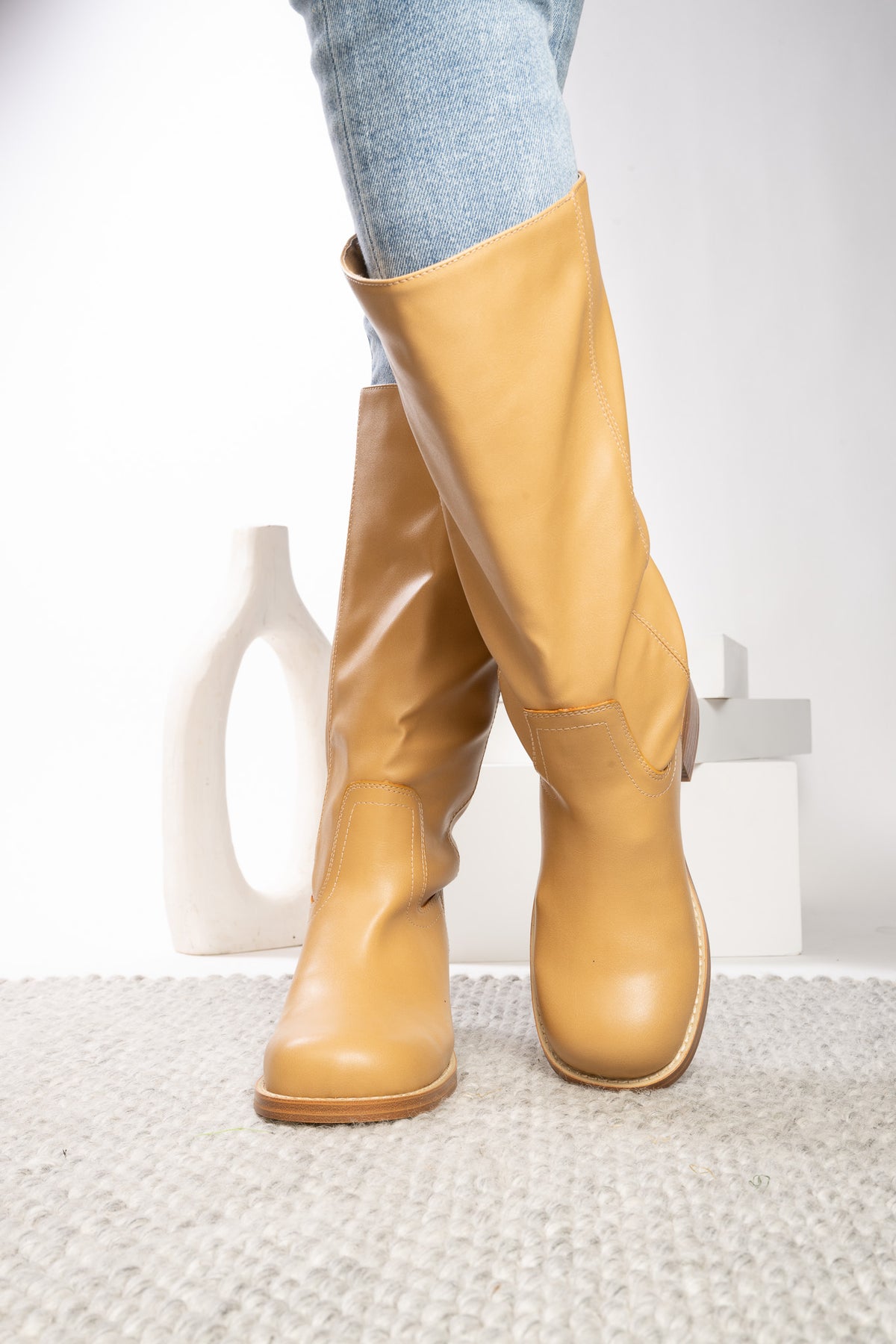 A pair of knee-high tan leather boots worn with light blue jeans, standing on a textured light gray rug with a minimalist white vase and block in the background.