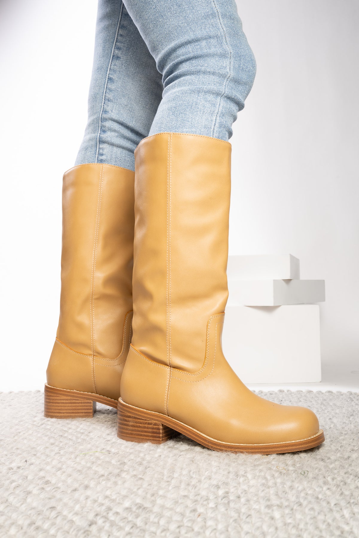 A pair of tan leather knee-high boots with stacked block heels, shown on a model wearing light-wash blue jeans against a white studio background.