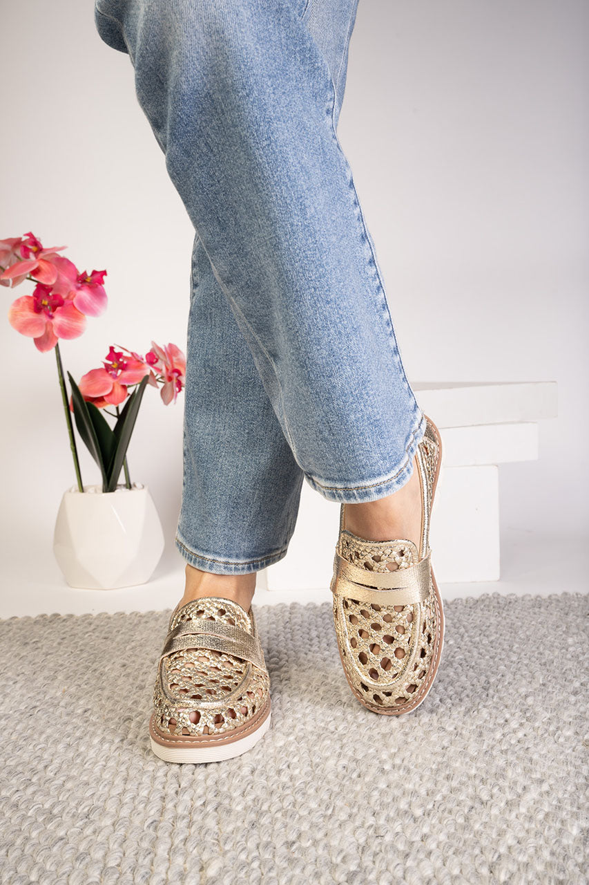 Person wearing Gold cut-out style loafers shoes and jeans with a white background and flowers.