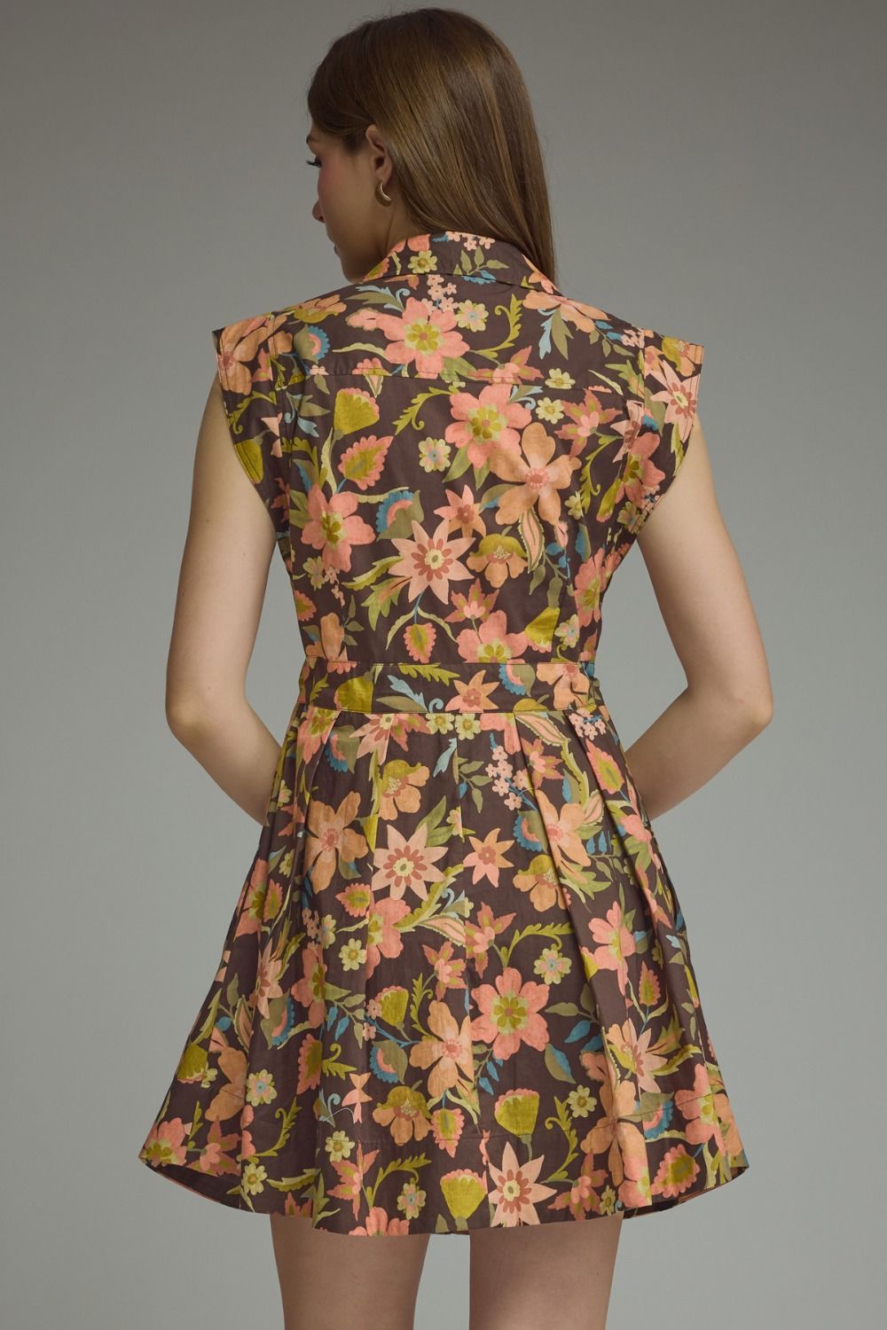 A woman shown from the back wearing a sleeveless, belted mini dress with a dense floral print on a dark brown background. The print features pink, peach, and yellow flowers with green leaves.
