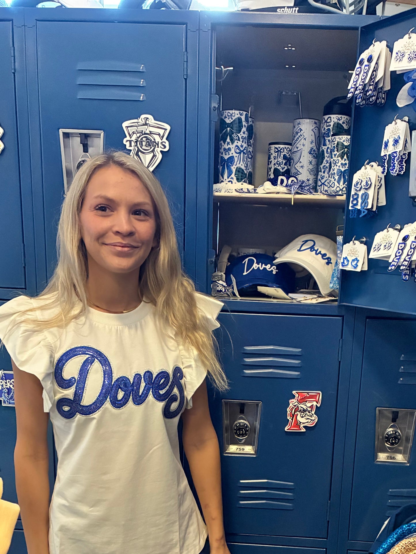 A young woman with blonde hair stands in front of blue school lockers wearing a white shirt with 'Doves' written in blue script across the chest. The locker is filled with Doves merchandise.