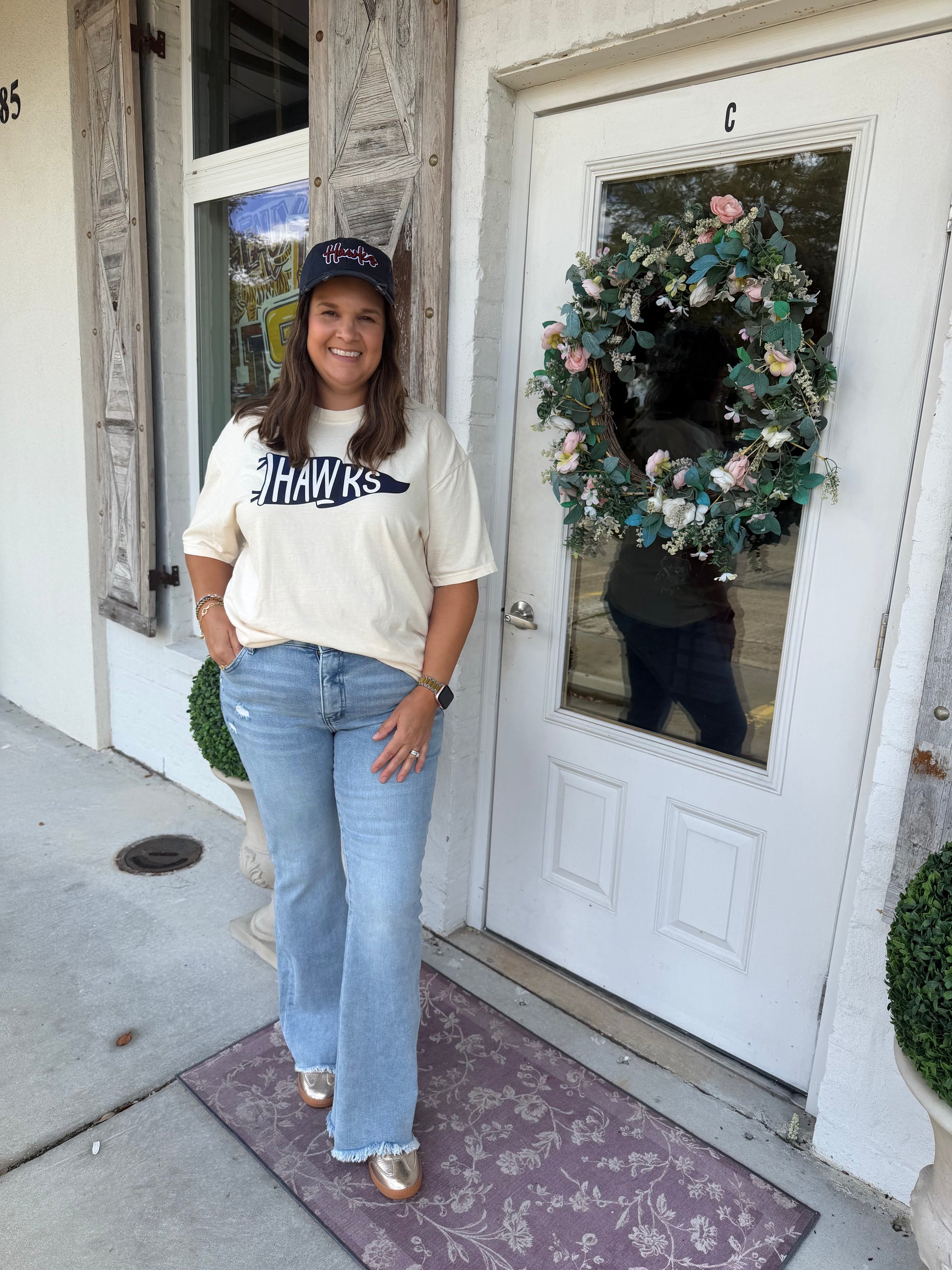 A woman wearing a cream graphic t-shirt with 'HAWKS' printed on the front, light blue flared jeans, and a Hawks navy baseball cap. 