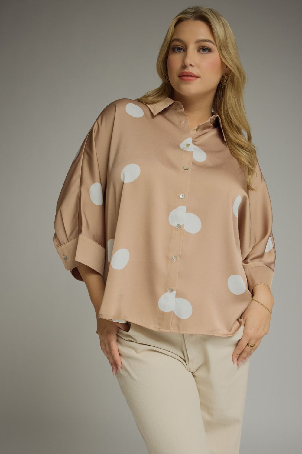 Woman wearing a beige blouse with white polka dots and beige pants against a gray background