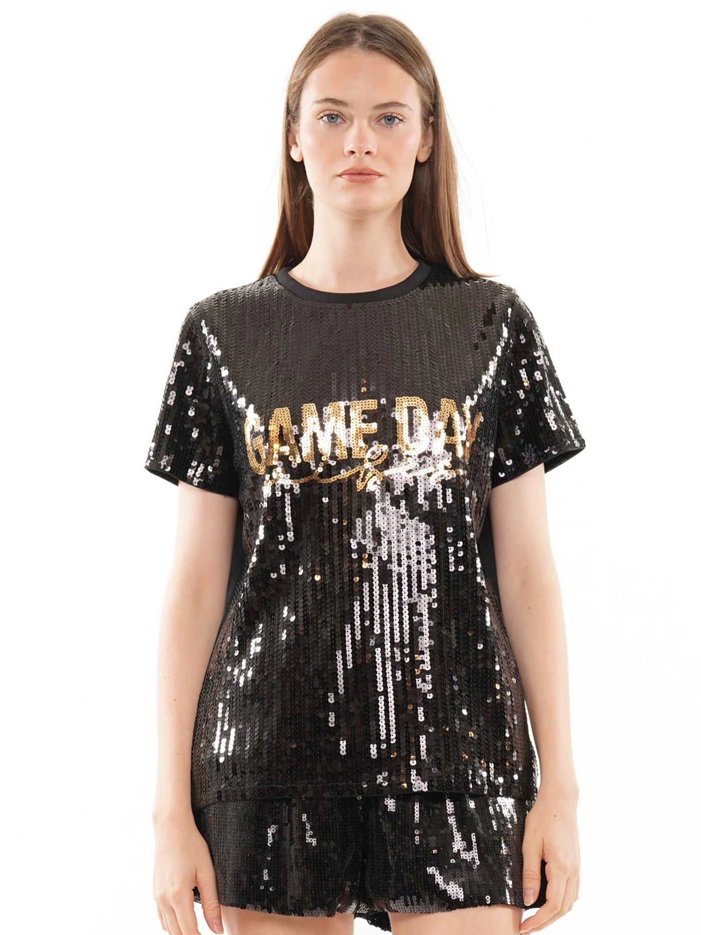 A woman modeling a black sequined t-shirt dress with 'GAME DAY' vibes printed across the chest in gold sequins, set against a plain white background.