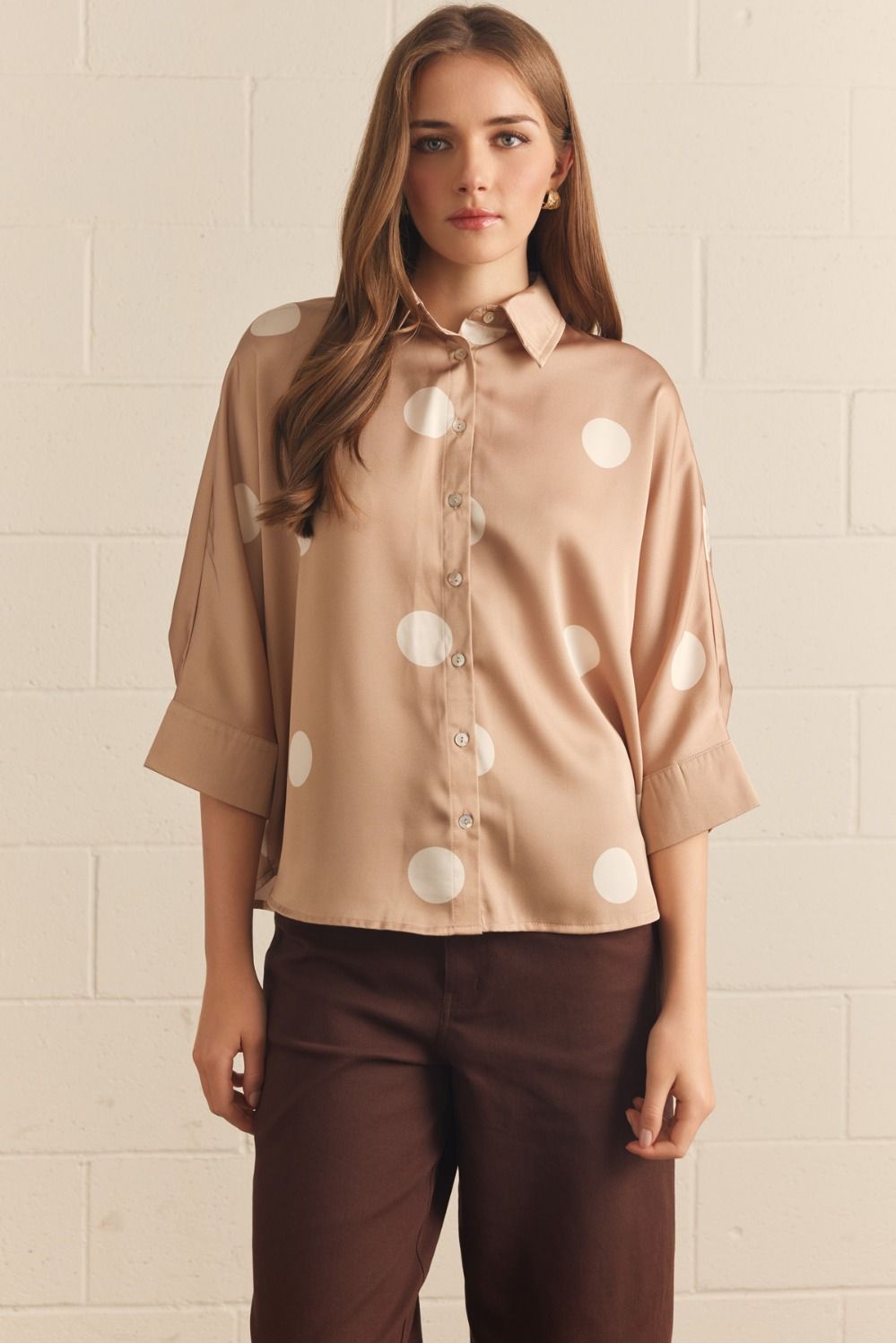 Woman wearing a beige blouse with white polka dots and brown pants against a beige wall.