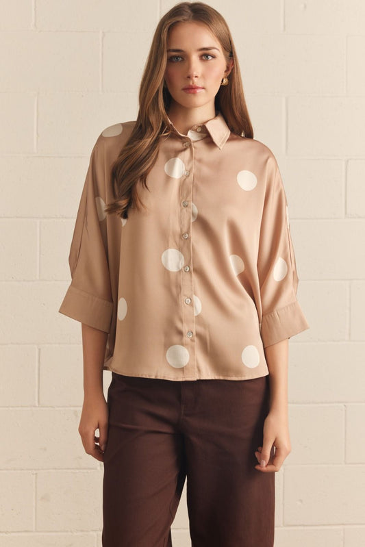Woman wearing a beige blouse with white polka dots and brown pants against a beige wall.