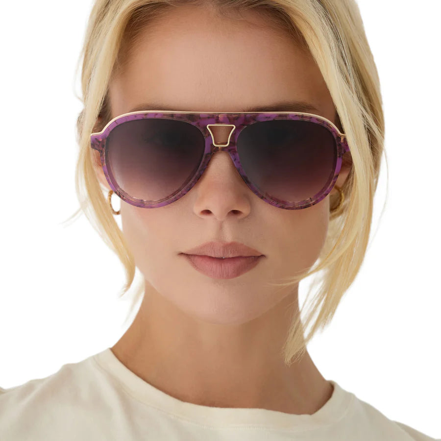 A close-up of a blonde model wearing aviator-style sunglasses with a purple tortoise design frame and a gold metal bridge detail, set against a plain white background.