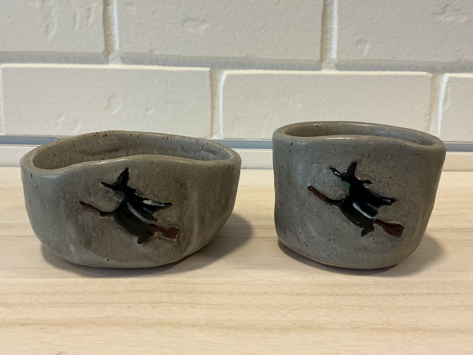 Two small handmade pottery candles with a gray glaze, featuring a black and brown witch riding a broomstick motif, set on a light wood surface against a white brick wall.