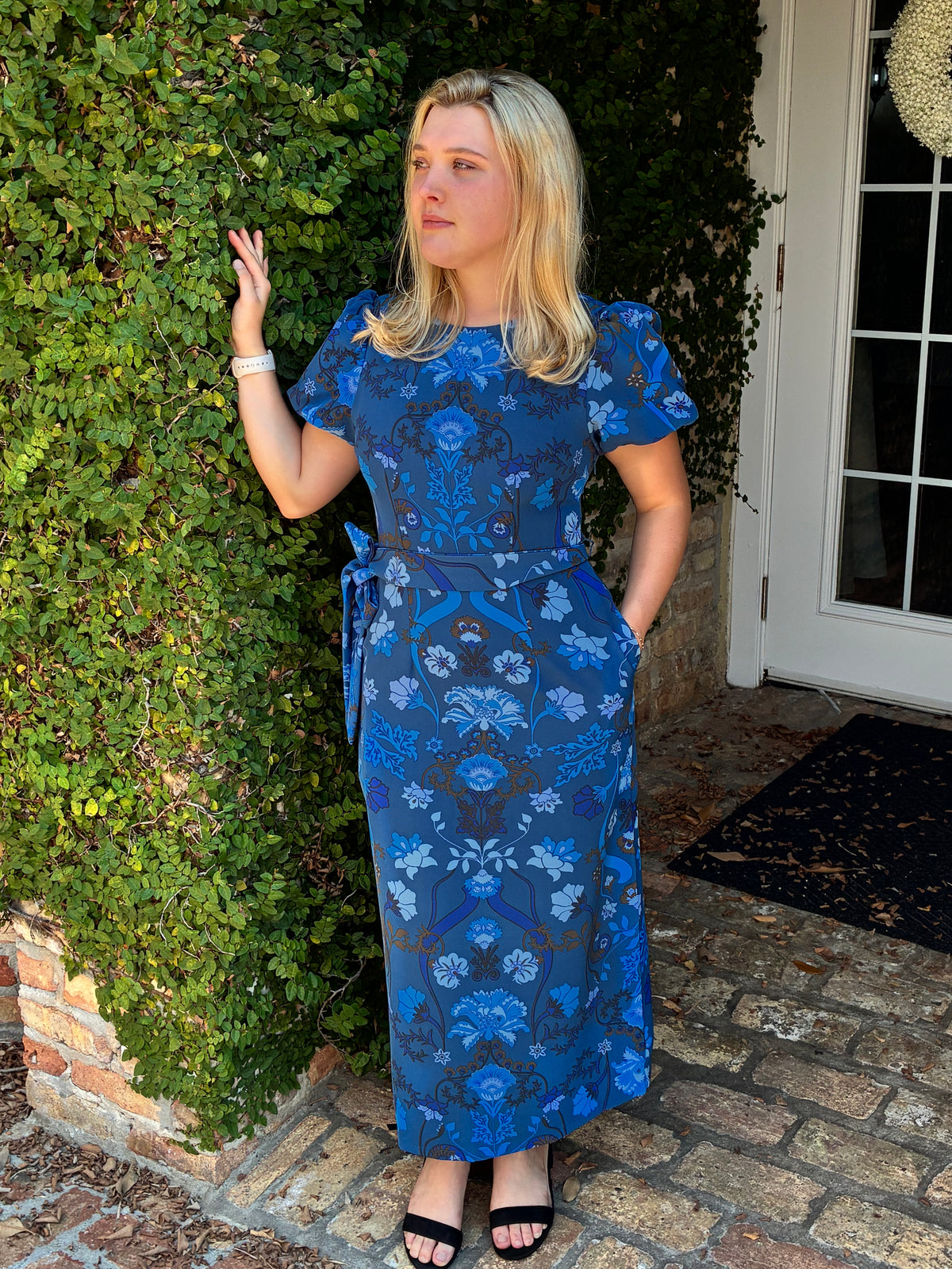 A woman wearing a blue patterned midi dress with short puff sleeves and a matching tie belt, standing outdoors next to a green hedge and brick patio.
