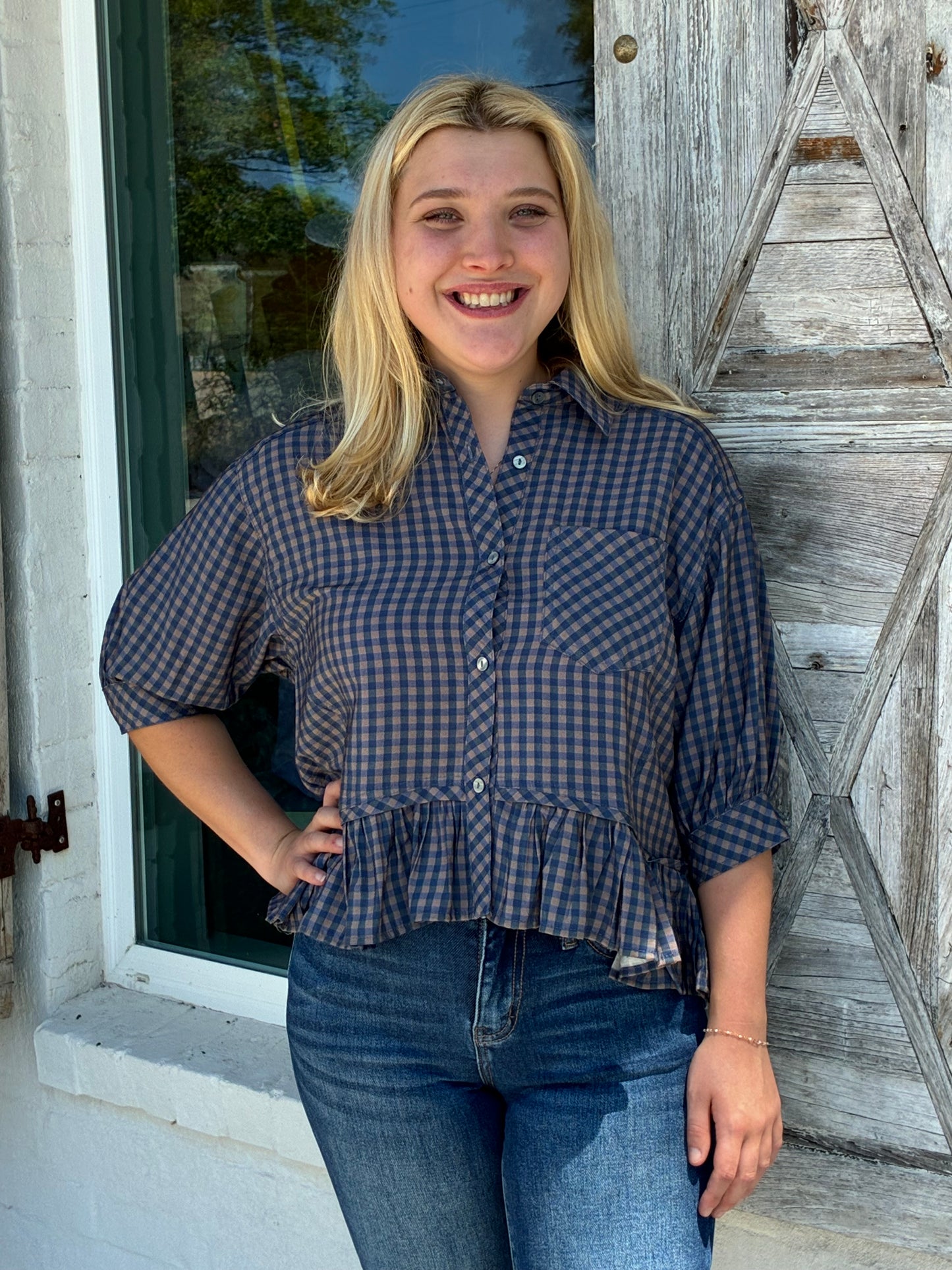 A smiling blonde woman wearing a blue and brown gingham button-up shirt with puff sleeves and a ruffled hem, paired with blue jeans, standing outdoors in front of a rustic wooden door.