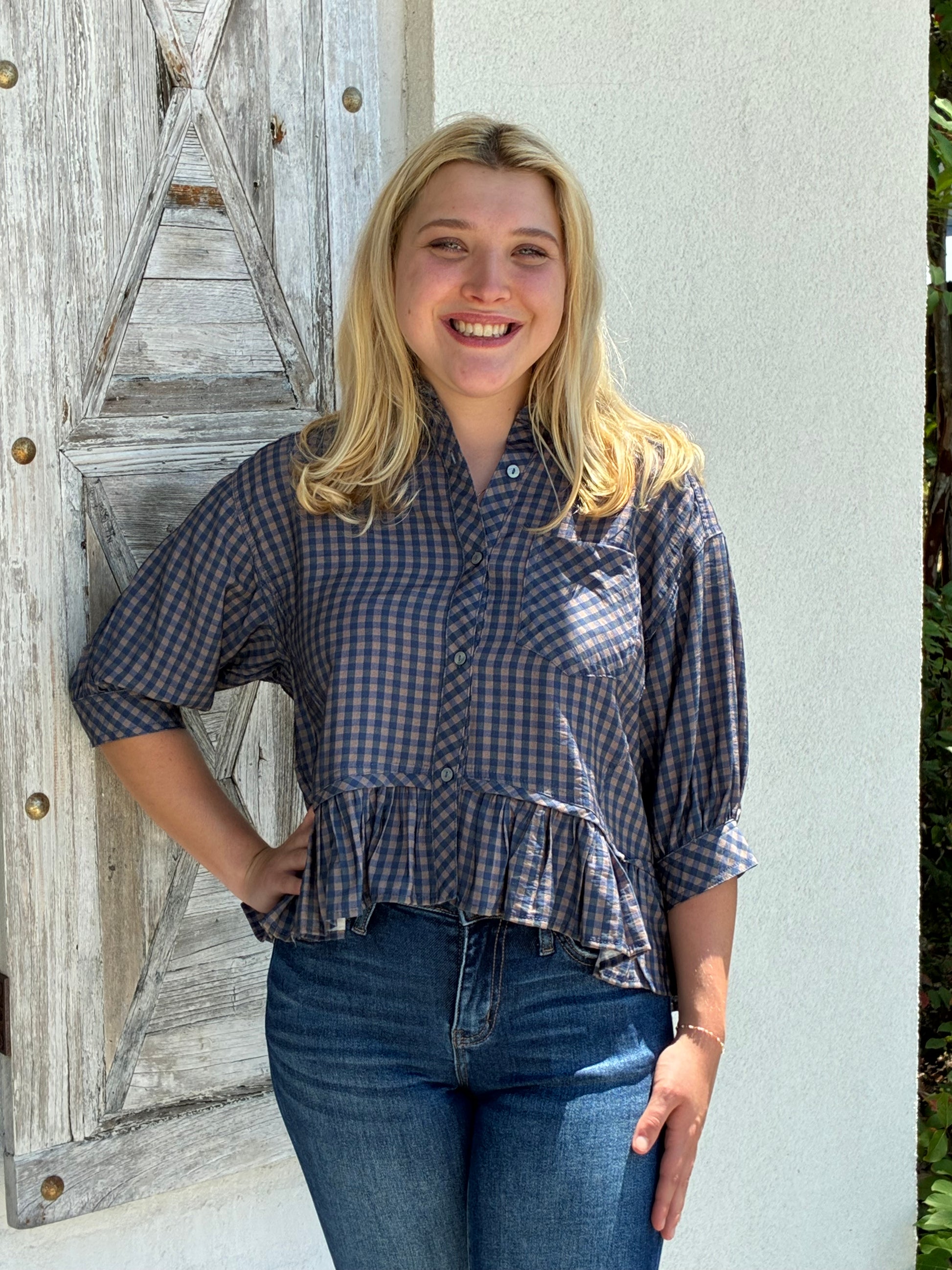 A smiling blonde woman wearing a blue and gray gingham button-up blouse with a ruffled hem and three-quarter sleeves, paired with blue jeans, standing outdoors against a white wall and a weathered wooden door.