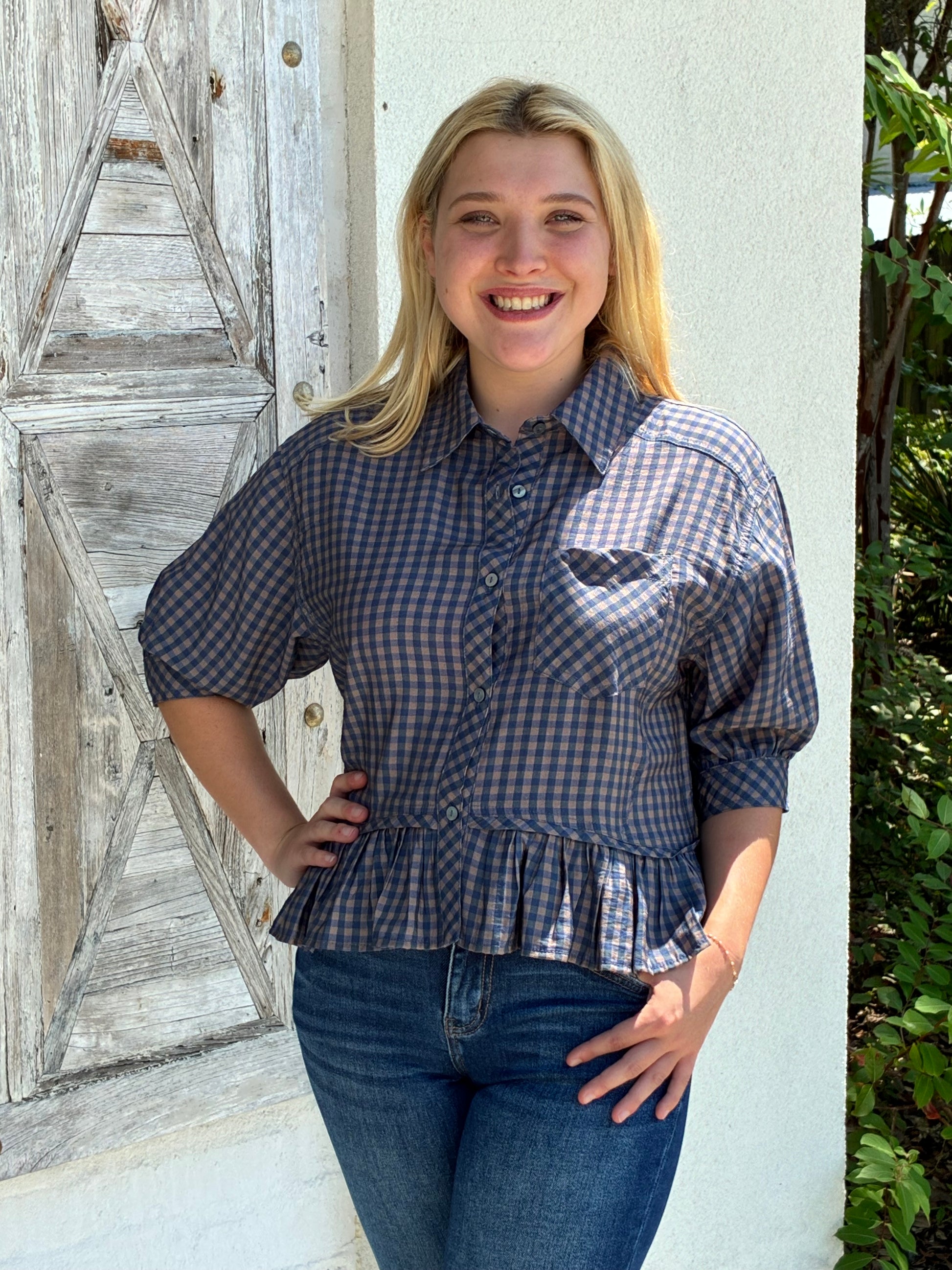 A smiling blonde woman wearing a blue and gray gingham check blouse with a ruffled hem and blue jeans, standing outdoors against a white wall and weathered wooden door.