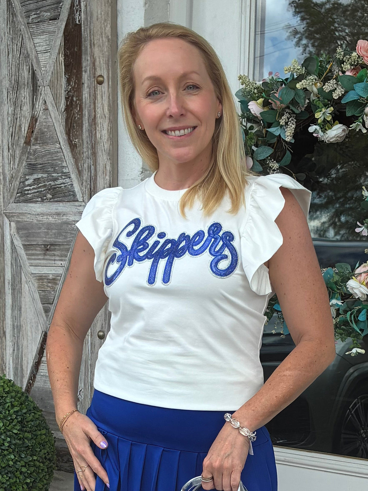 A woman stands on a storefront porch wearing a white ruffle-sleeve t-shirt with 'Slippers' in blue script, a royal blue pleated mini skirt, metallic silver sneakers, and holding a small silver handbag.