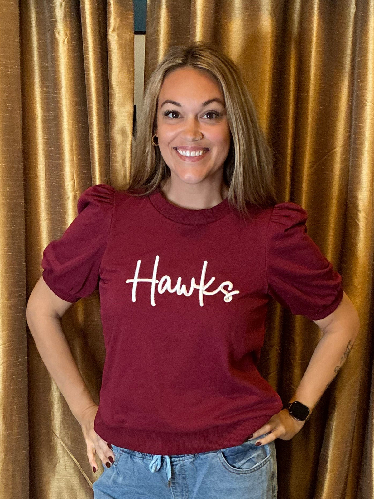 A woman stands in front of gold curtains wearing a burgundy t-shirt with 'Hawks' written in white script across the chest, paired with distressed light blue jeans and brown sandals.