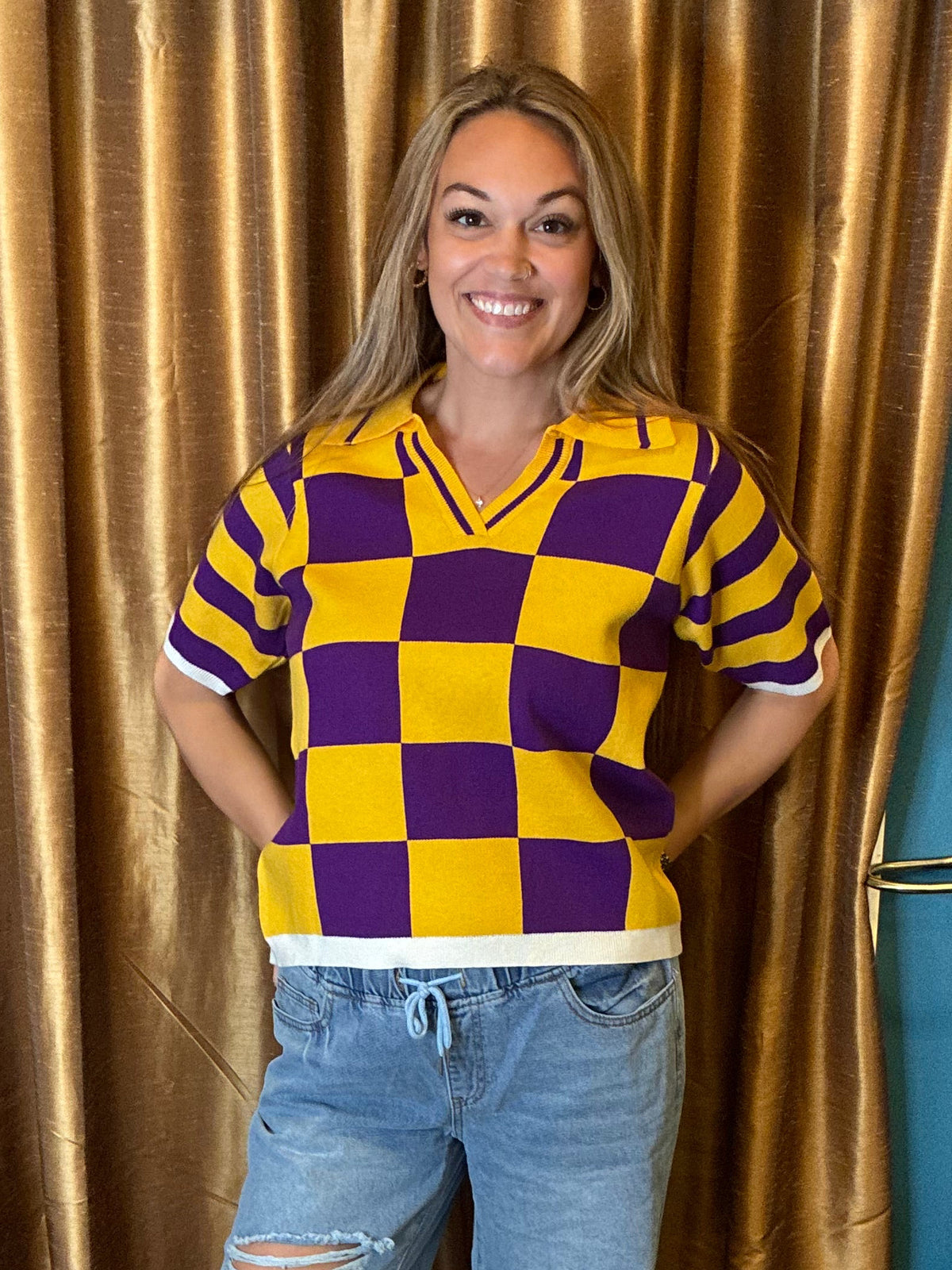 A woman stands in a room wearing a yellow and purple checkered knit sweater, distressed light-wash jeans, and black sandals. She is positioned in front of a large gold curtain with a blue wall behind it.