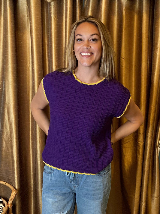 A woman stands in front of gold curtains wearing a purple knit top with yellow trim and light blue ripped jeans, in a room with a blue wall and a small clothing rack.