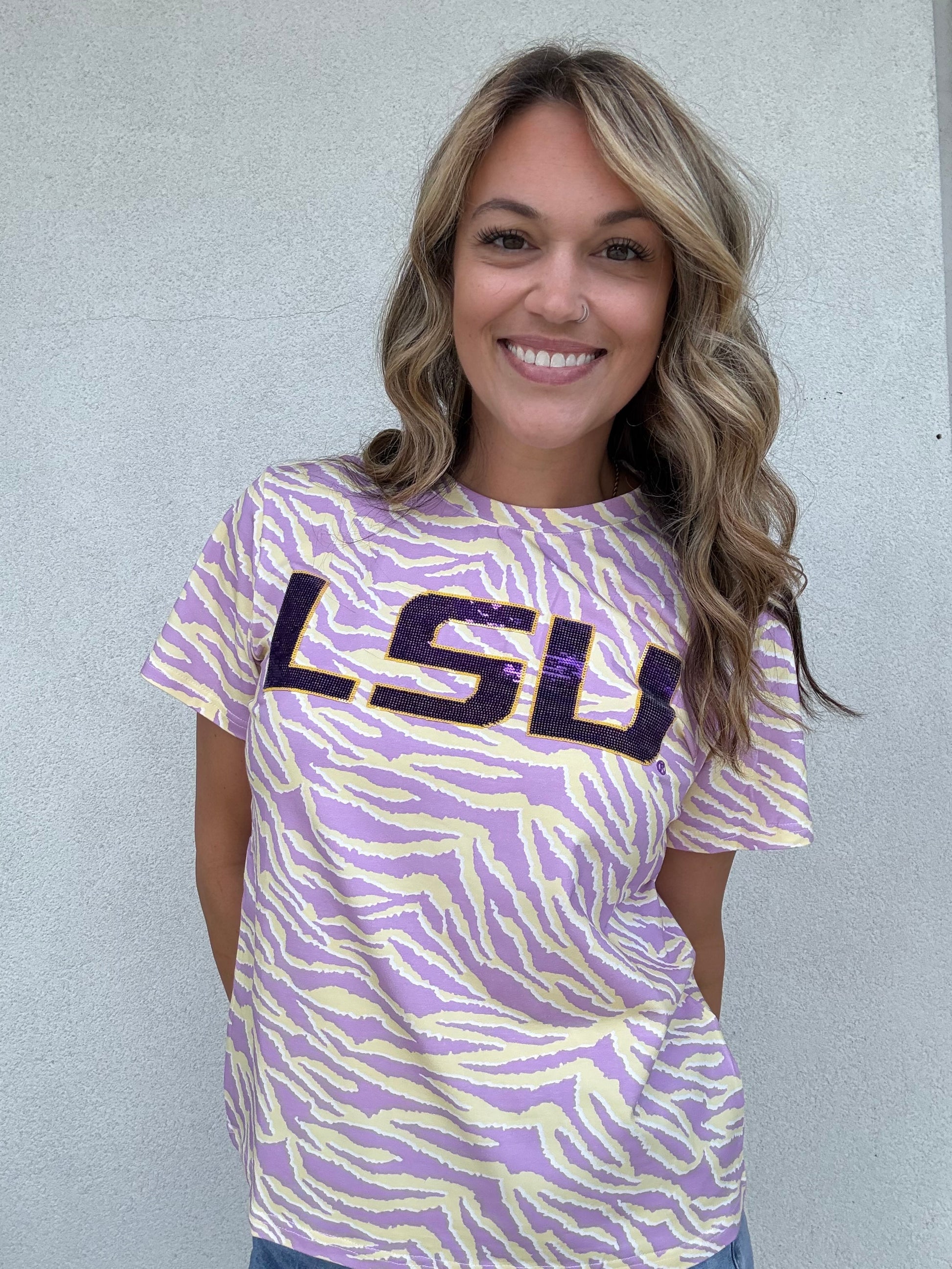 A smiling woman with blonde-highlighted hair wearing a purple and yellow tiger-print t-shirt with 'LSU' printed across the chest, standing against a light gray stucco wall.