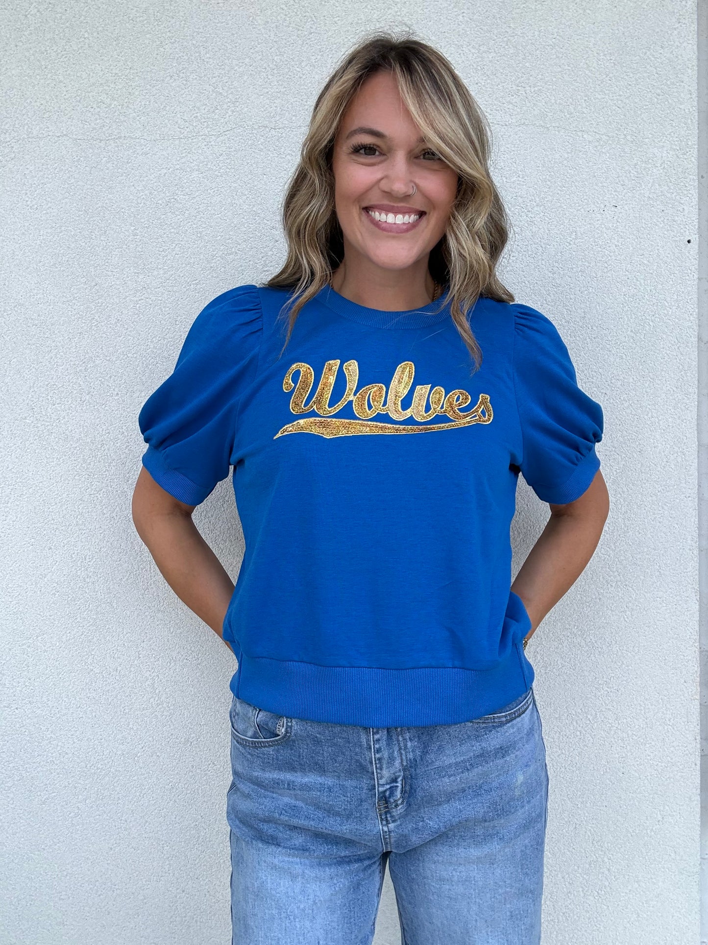 A woman smiling and wearing a royal blue crewneck sweatshirt with 'Wolves' written across the chest in gold sequin lettering, paired with light-wash blue jeans, standing against a light gray textured wall.