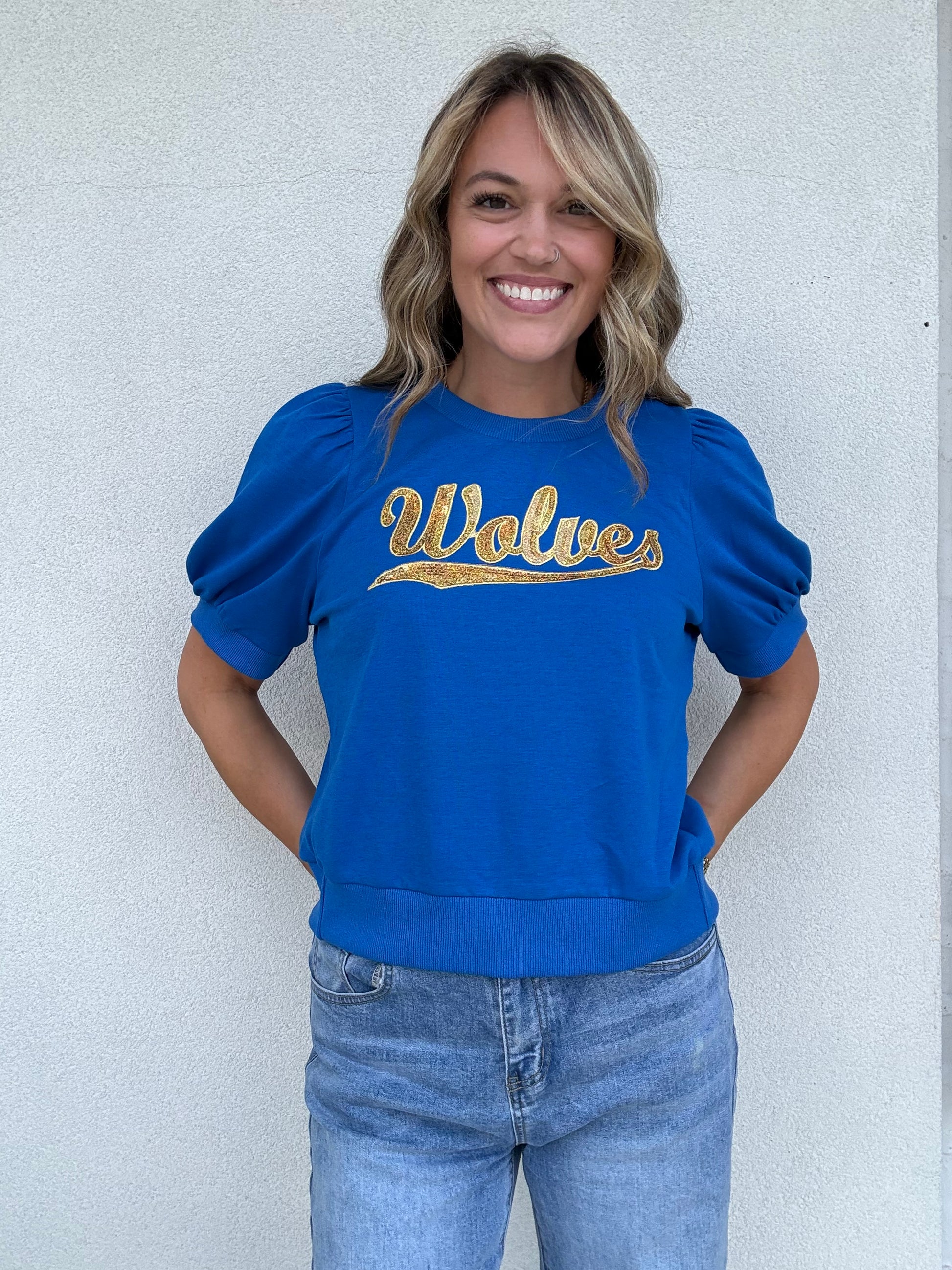 A woman smiling and wearing a royal blue crewneck sweatshirt with 'Wolves' written across the chest in gold sequin lettering, paired with light-wash blue jeans, standing against a light gray textured wall.
