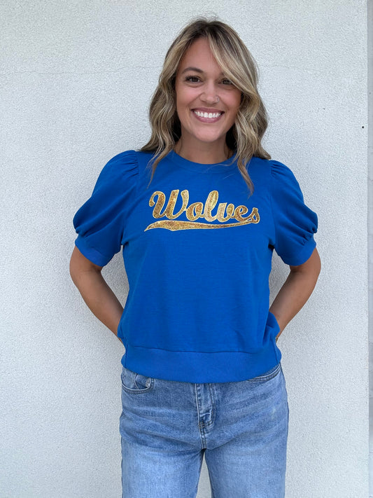 A woman smiling and wearing a royal blue crewneck sweatshirt with 'Wolves' written across the chest in gold sequin lettering, paired with light-wash blue jeans, standing against a light gray textured wall.