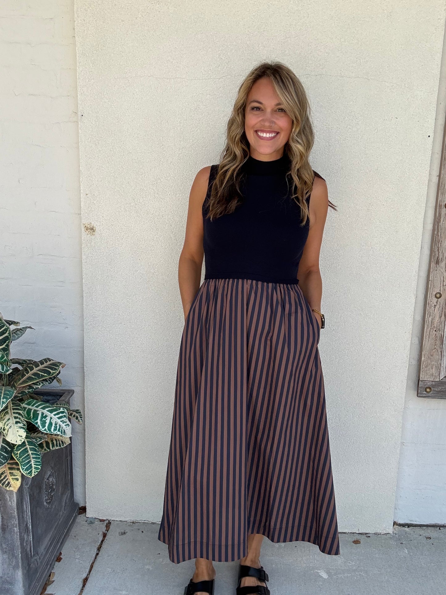 A woman stands outdoors against a white stucco wall wearing a sleeveless mock-neck midi dress with a solid navy top and vertical brown and navy stripes on the skirt.