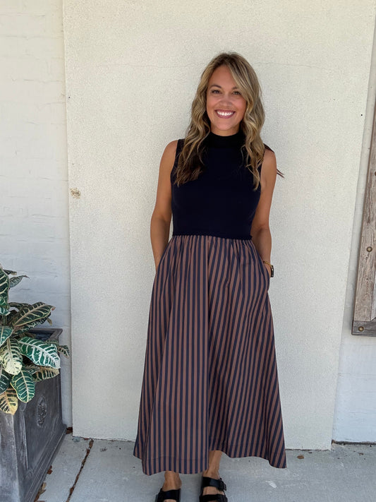 A woman stands outdoors against a white stucco wall wearing a sleeveless mock-neck midi dress with a solid navy top and vertical brown and navy stripes on the skirt.