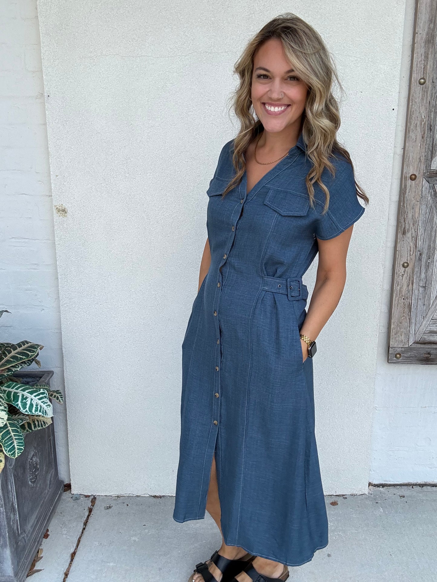 A woman wearing a blue denim  print midi dress with short sleeves, a button-up front, and a matching belt, standing against a white stucco wall.
