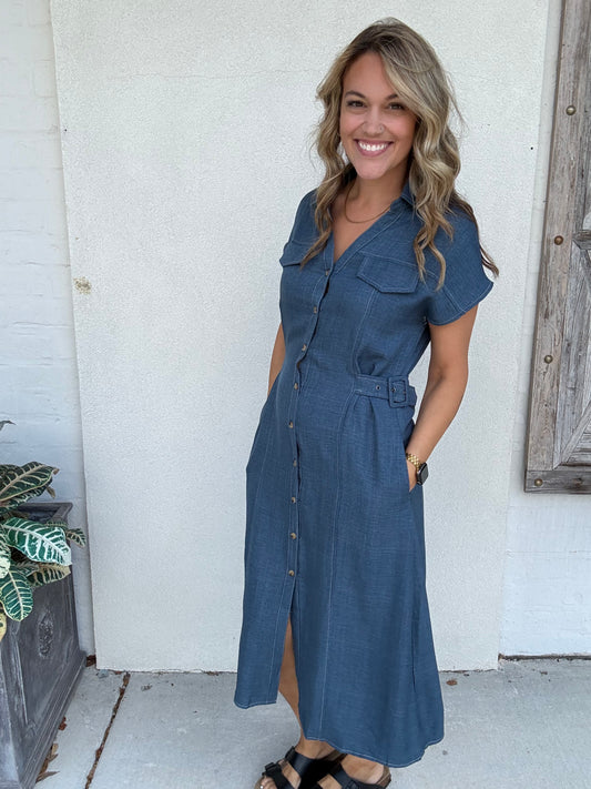 A woman wearing a blue denim  print midi dress with short sleeves, a button-up front, and a matching belt, standing against a white stucco wall.