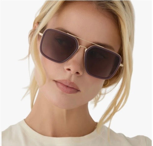 A close-up of a blonde woman wearing oversized square sunglasses with dark tinted lenses and thin gold metal frames, set against a clean white background.