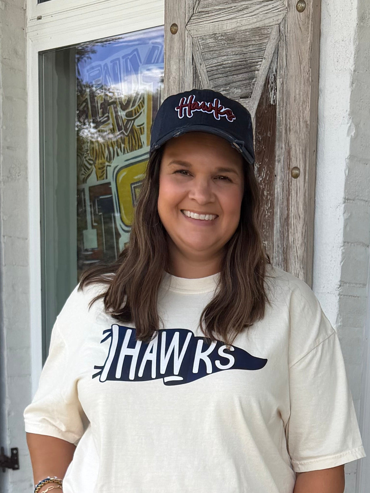 A woman stands on a purple patterned doormat in front of a white door with a floral wreath, wearing a cream graphic t-shirt with 'Hawks' printed on the chest, light blue flared jeans, and a Navy baseball cap with Hawks embroidery 