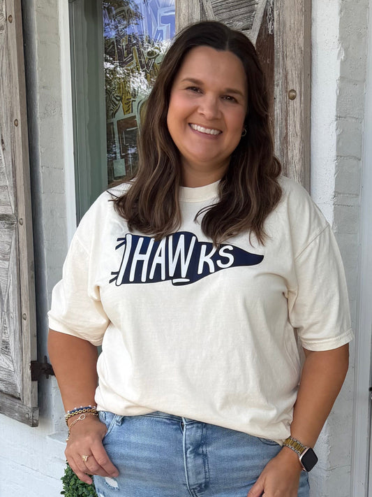 A woman stands on a porch in front of a white door with a floral wreath, wearing a cream graphic t-shirt with 'HAWKS' printed on the front in Navy , light blue flared jeans, and gold shoes.