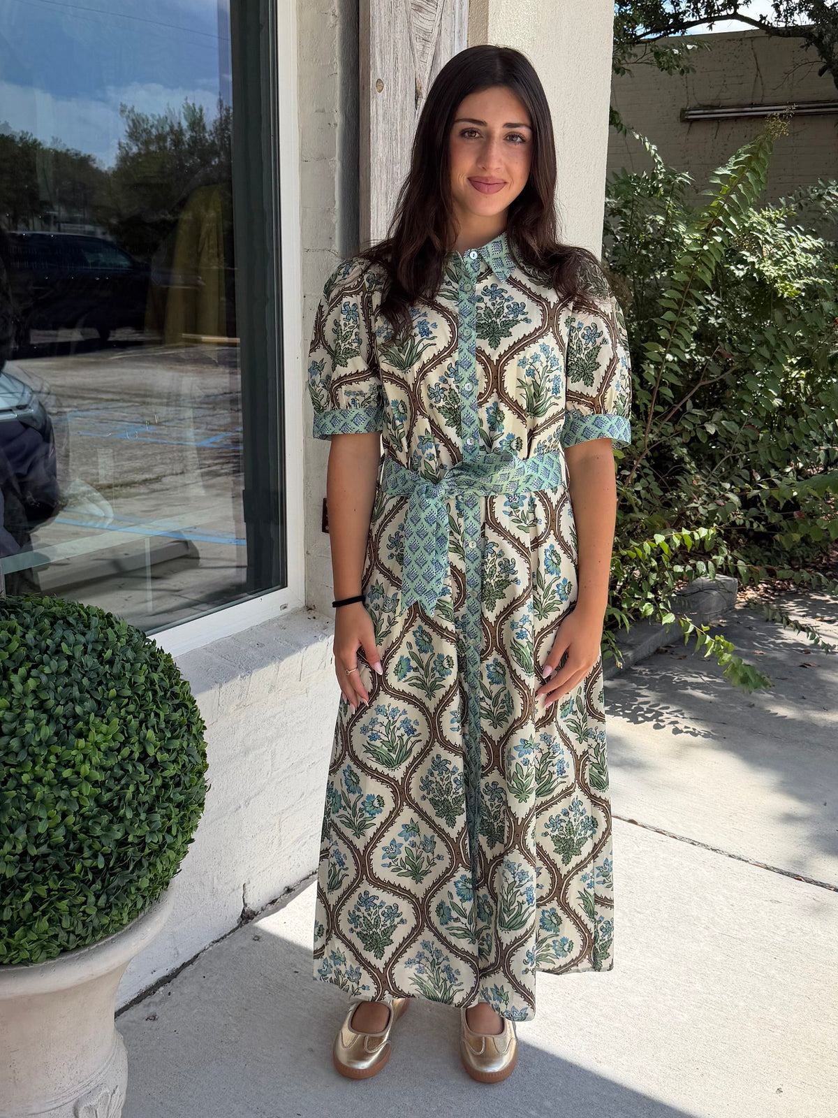 A woman stands outdoors on a porch wearing a short-sleeve midi dress with a green, brown, blue and beige geometric botanical . The dress is button up with a tie.