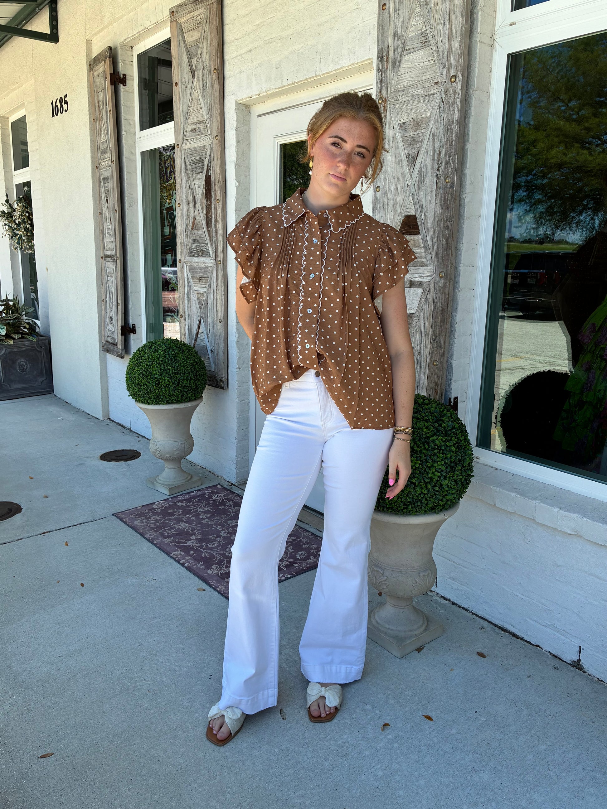 A woman posing outdoors on a concrete walkway in front of a white building, wearing a brown short-sleeve button-up shirt with a white polka-dot pattern and white wide-leg pants.