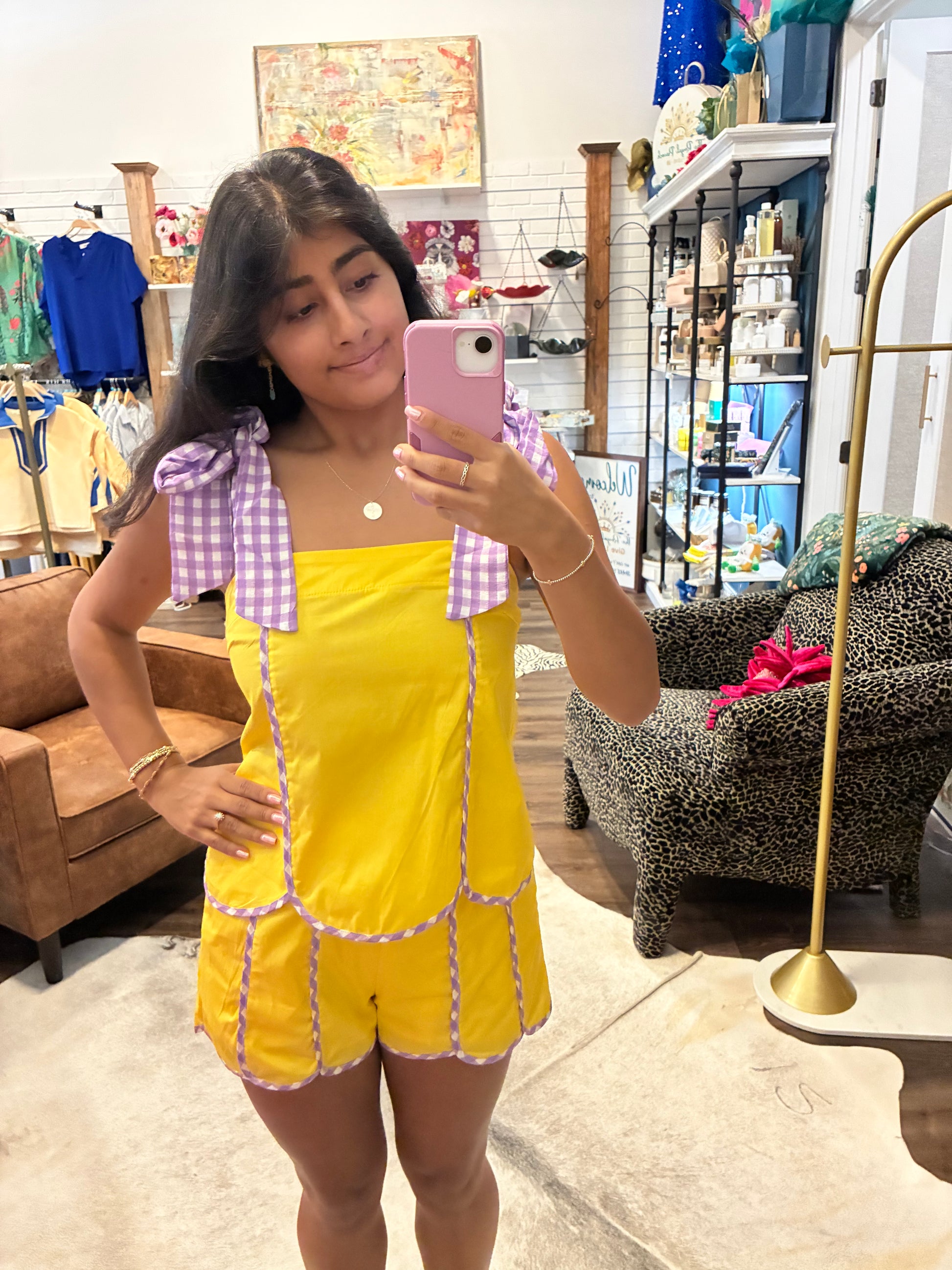 A woman takes a mirror selfie in a boutique while wearing a bright yellow romper with lavender gingham bow details on the shoulders and trim along the front seams.