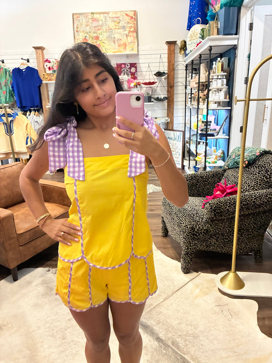 A woman takes a mirror selfie in a boutique while wearing a bright yellow romper with lavender gingham bow details on the shoulders and trim along the front seams.