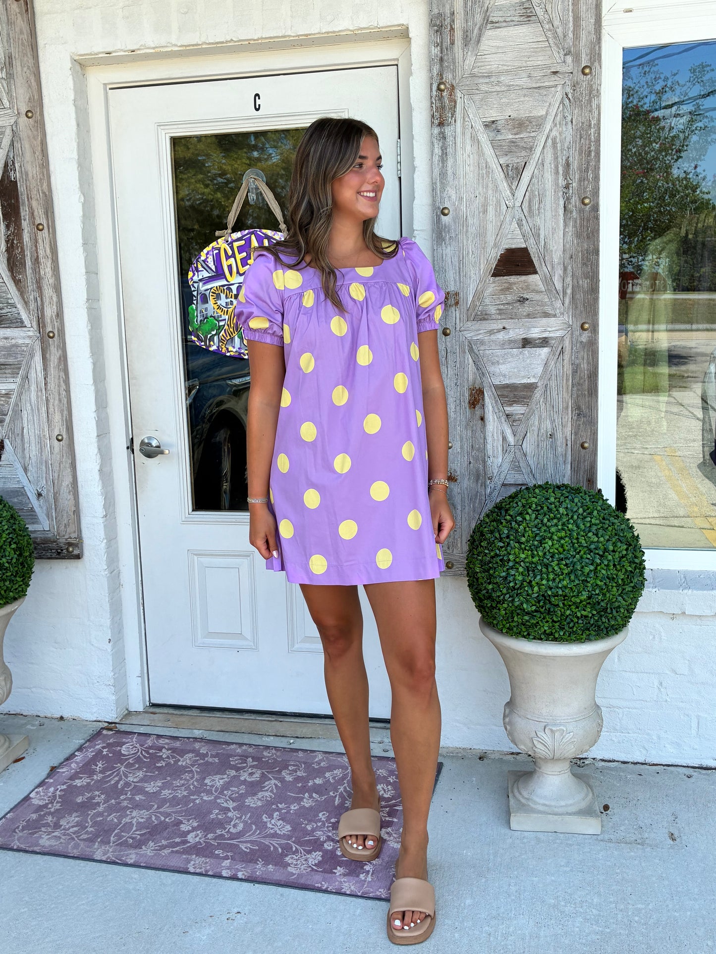 A woman wearing a lavender purple short-sleeve mini dress with yellow polka dots, standing on a porch in front of a white door with rustic wooden shutters.