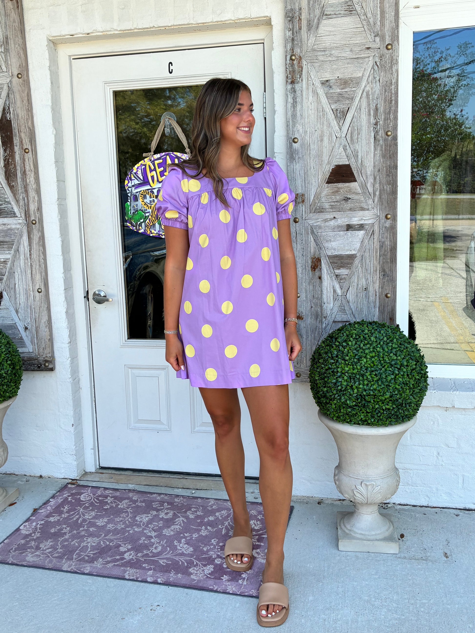 A woman wearing a lavender purple short-sleeve mini dress with yellow polka dots, standing on a porch in front of a white door with rustic wooden shutters.