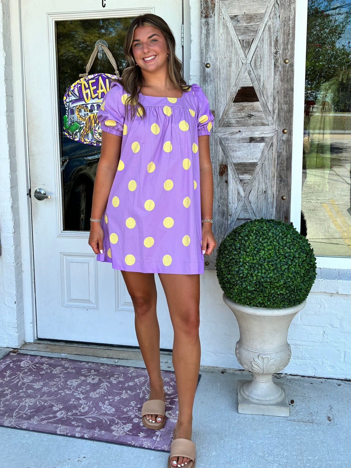 A young woman wearing a purple short-sleeve dress with yellow polka dots, standing on a white porch in front of a door with a wooden shutters and a potted topiary.