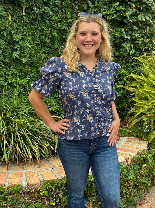 A young woman with blonde hair smiling while wearing a navy blue floral short-sleeve blouse and blue jeans, standing outdoors in a garden setting with green plants and a brick path.