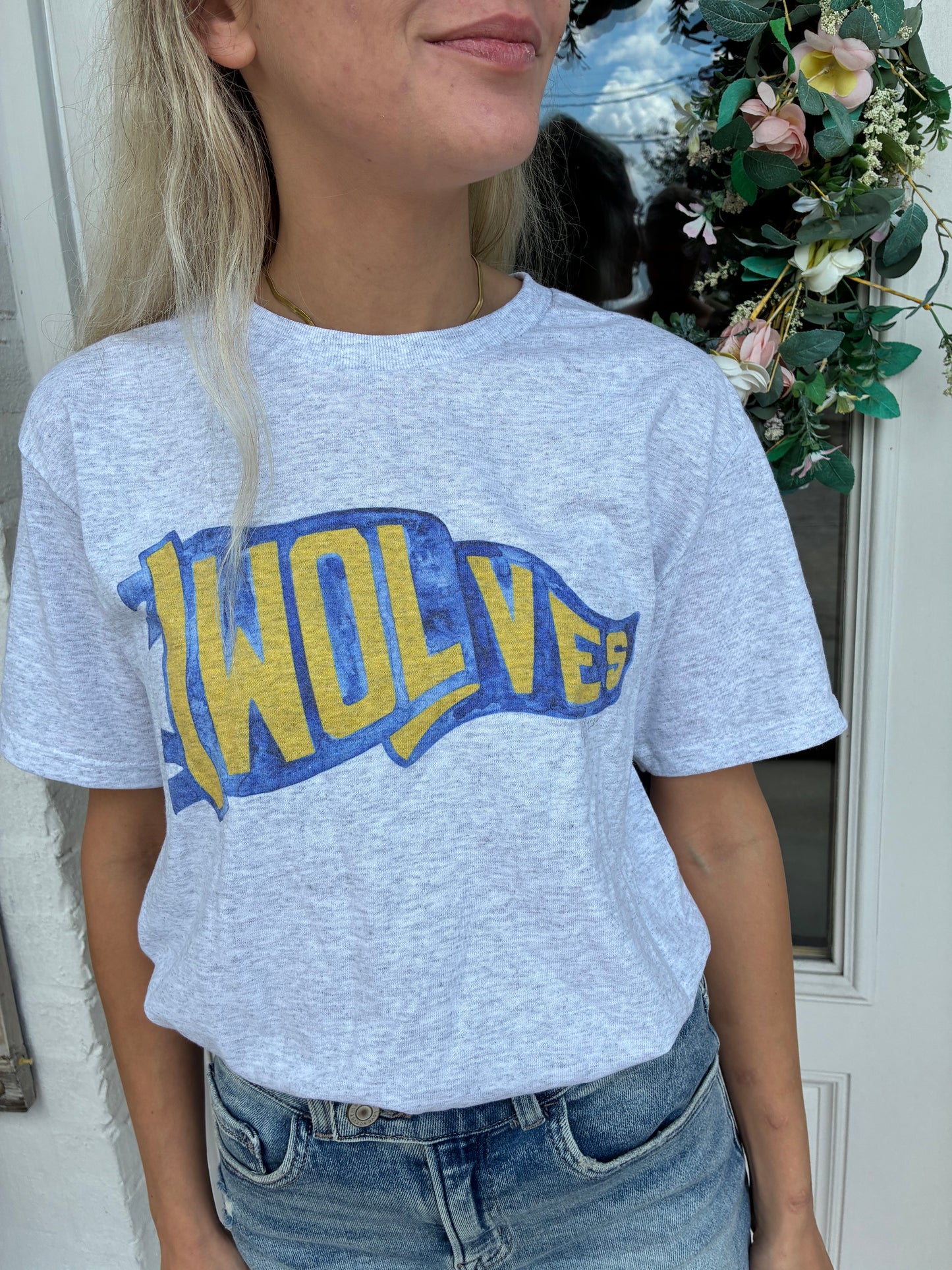 A woman wearing a light heather gray t-shirt with a blue and yellow 'WOLVES' flag pennant graphic, paired with blue jeans, standing in front of a white door with a floral wreath.