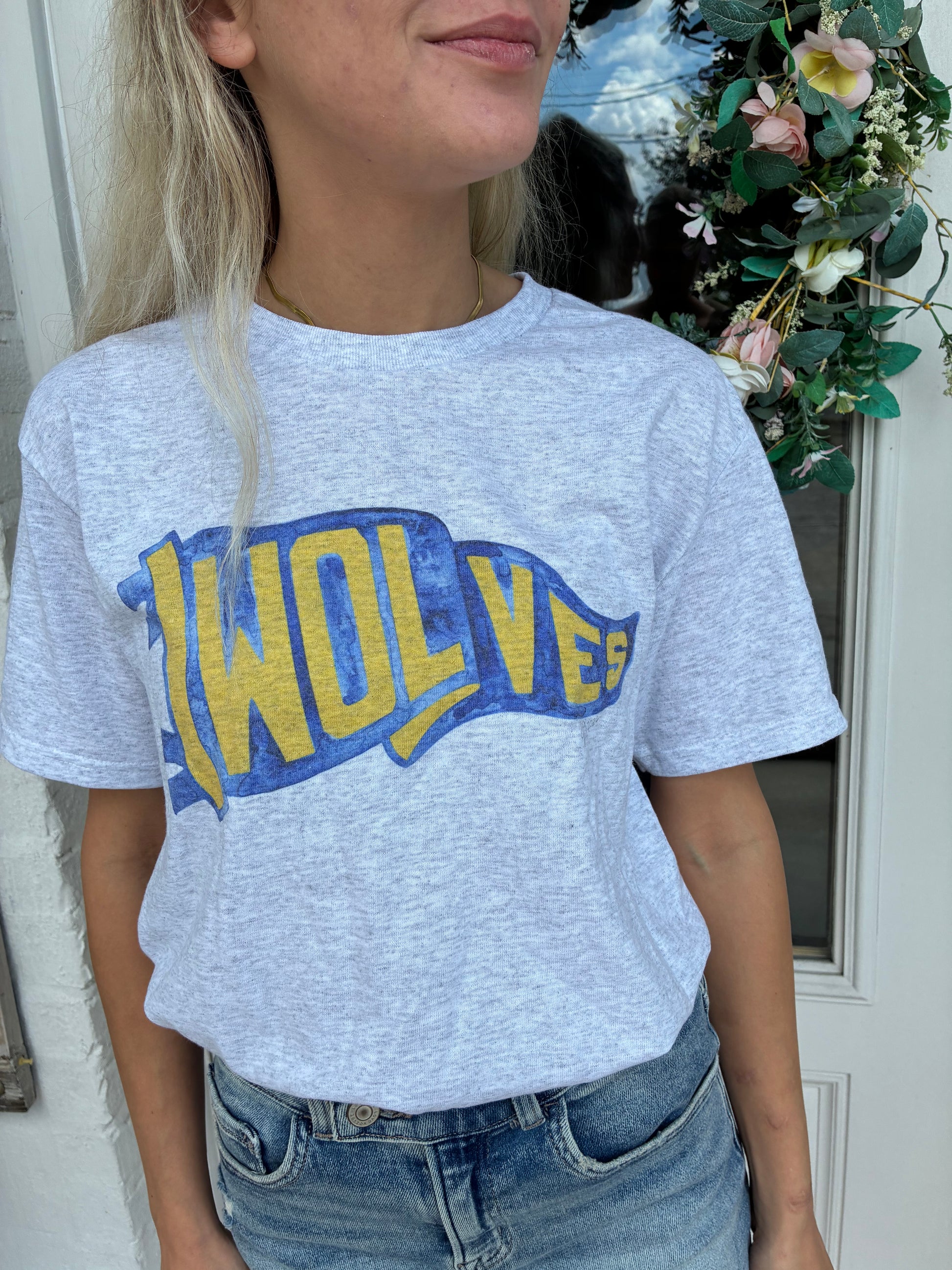 A woman wearing a light heather gray t-shirt with a blue and yellow 'WOLVES' flag pennant graphic, paired with blue jeans, standing in front of a white door with a floral wreath.