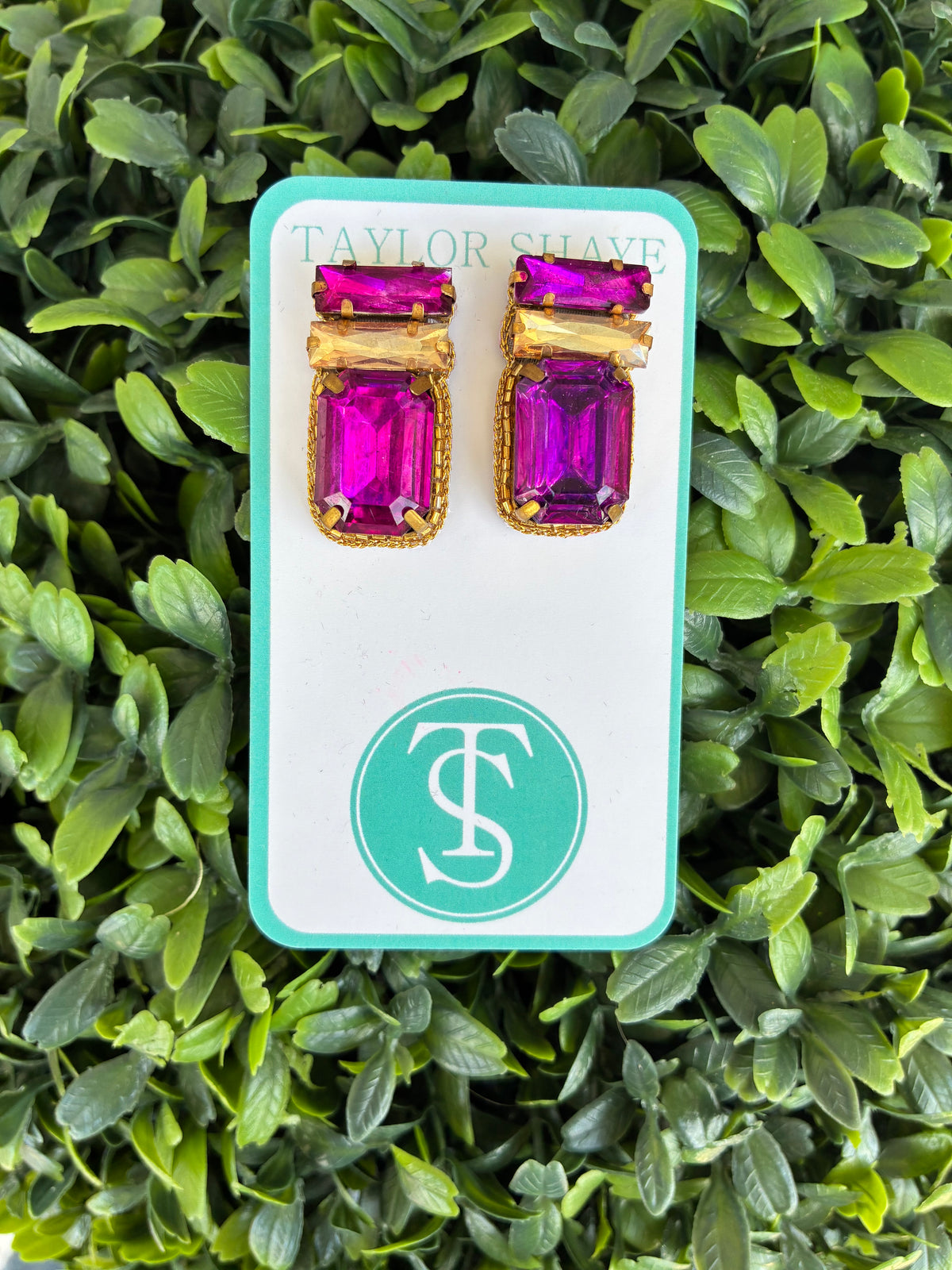 A pair of gold-toned stud earrings with rectangular purple gemstones, displayed on a white card with a teal border and the Taylor Shave logo, set against a background of green leaves.