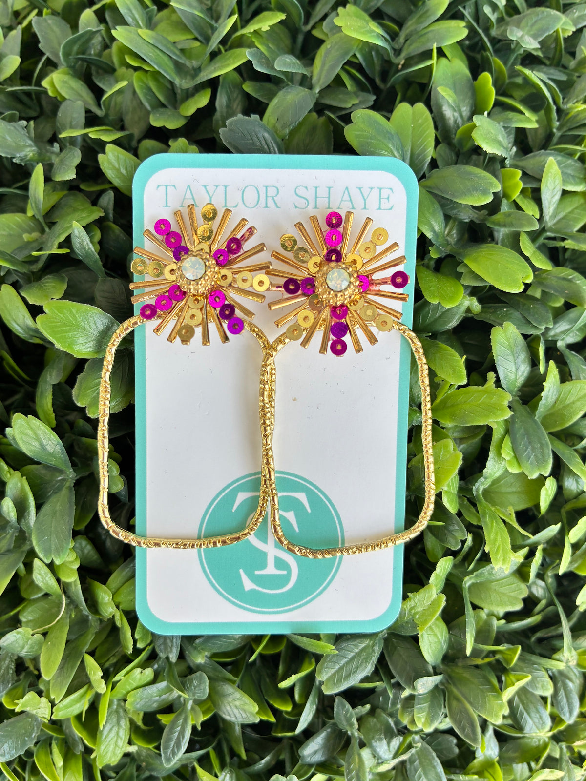 A pair of gold-toned starburst stud earrings with purple and gold rhinestone accents, displayed on a turquoise and white Taylor Shaye card against a background of green foliage.